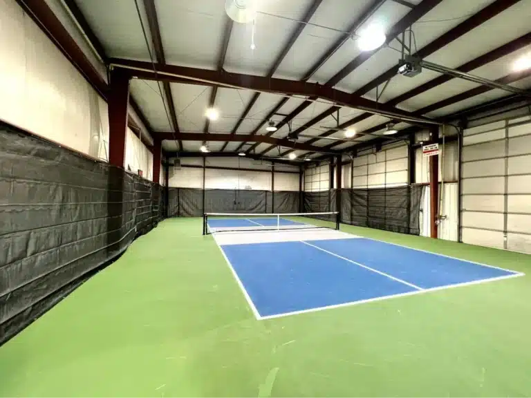 An indoor tennis court with blue and green surfaces, surrounded by tall, black netting. The ceiling is open with visible beams and lighting, creating the perfect environment for facility owners to consider adding indoor pickleball courts.