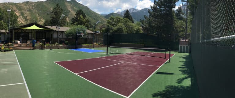 Outdoor sports court with basketball hoops, tennis net, and green and maroon surfaces. Surrounded by trees and mountains in the background, with houses and a covered area to the side.