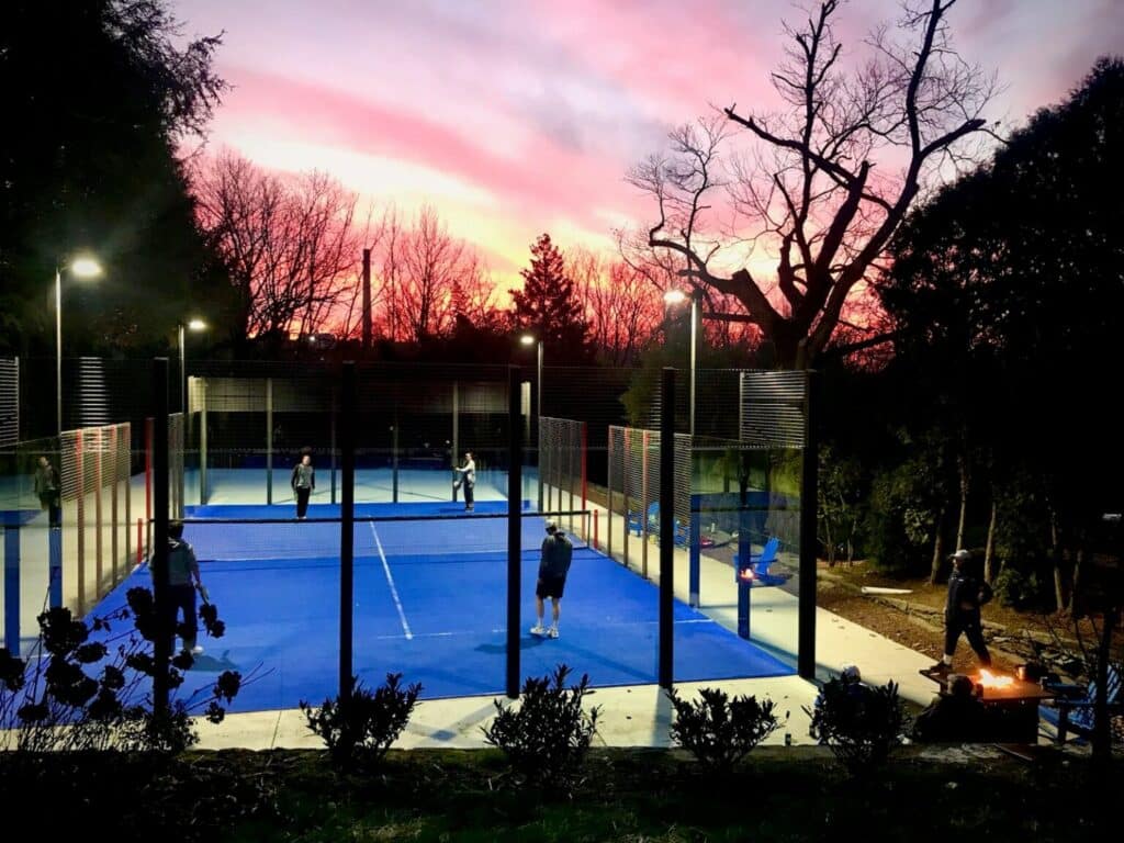 Four people engage in a friendly match on a vibrant blue padel court at dusk, framed by fencing and trees, while a colorful sunset paints the sky. It's an atmospheric backdrop reminiscent of the Series G's perfect alignment of elements.