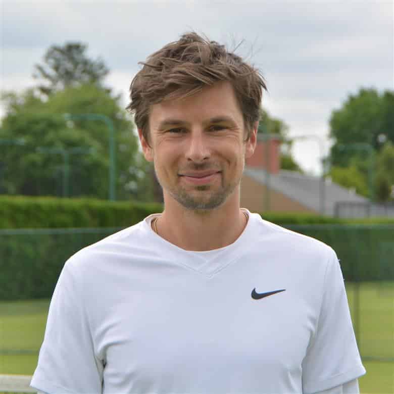 A person with short brown hair and a beard stands outdoors, wearing a white HCA Sports Nike shirt. Trees, greenery, and a building are visible in the background.