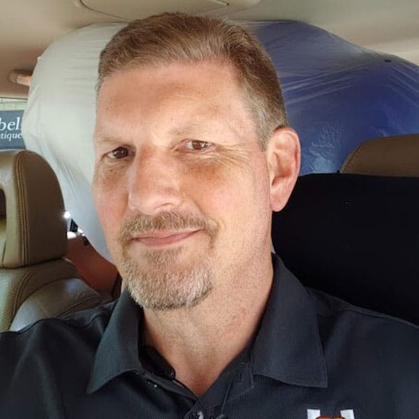 A man with short hair and a goatee is seated in a car, looking at the camera with a slight smile. He is wearing a dark-colored HCA Sports polo shirt.