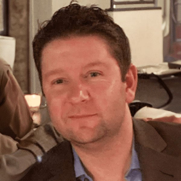 A man with short dark hair and a slight smile is wearing a blazer over a light blue shirt, seated indoors.