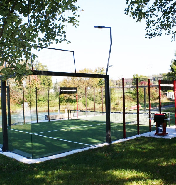 Residential padel court surrounded by netting and trees, with a bench and a maintenance machine nearby.