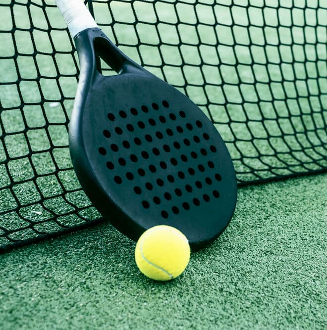 A black paddle and a yellow tennis ball rest against a net on a green paddle tennis court.